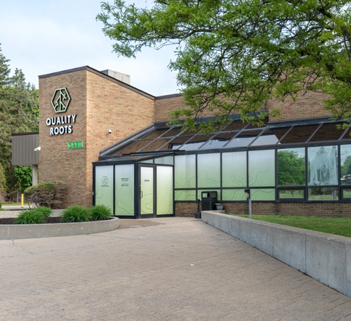 Mobile hero image of Quality Roots Owosso cannabis dispensary exterior with tree canopy, modern architecture, and welcoming entrance