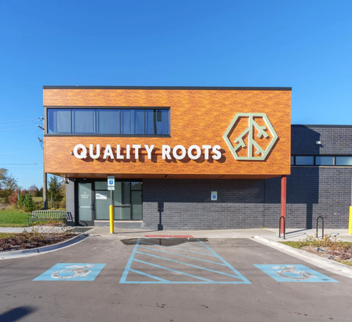 Mobile-optimized exterior view of Quality Roots Waterford recreational cannabis dispensary featuring modern gray building with large orange branded signage near M-59 in Oakland County Michigan